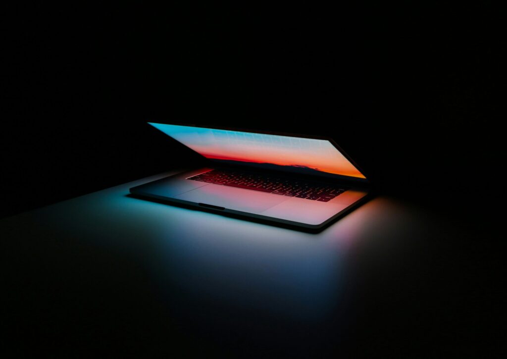 A slightly open laptop on a desk in a dark room, glowing with a vibrant sunset screen display that casts colorful light onto the surface below.